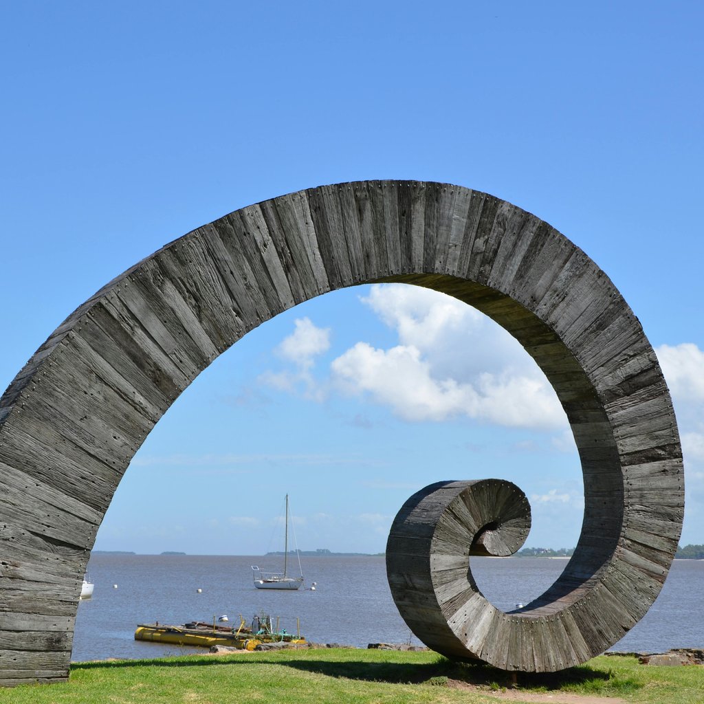 A spiral sculpture by the sea in Colonia Del Sacramento with sailboats in the background under a clear blue sky.