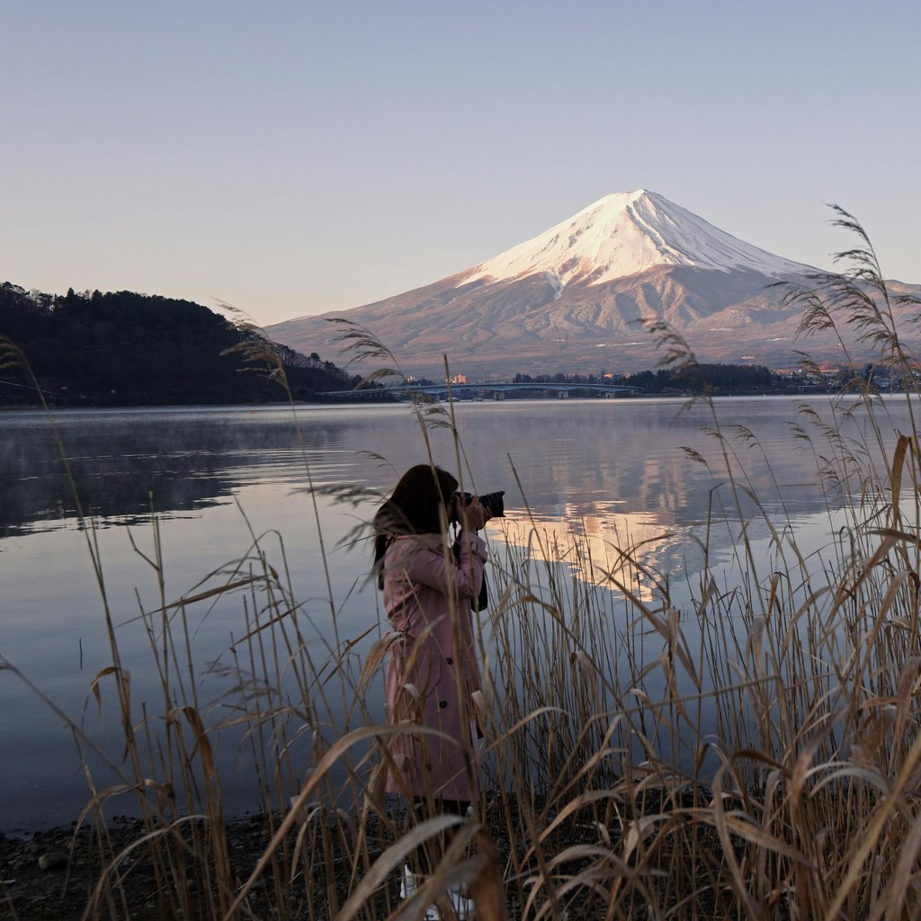 A woman captures the scenic beauty of Mount Fuji reflecting on a lake at sunset.