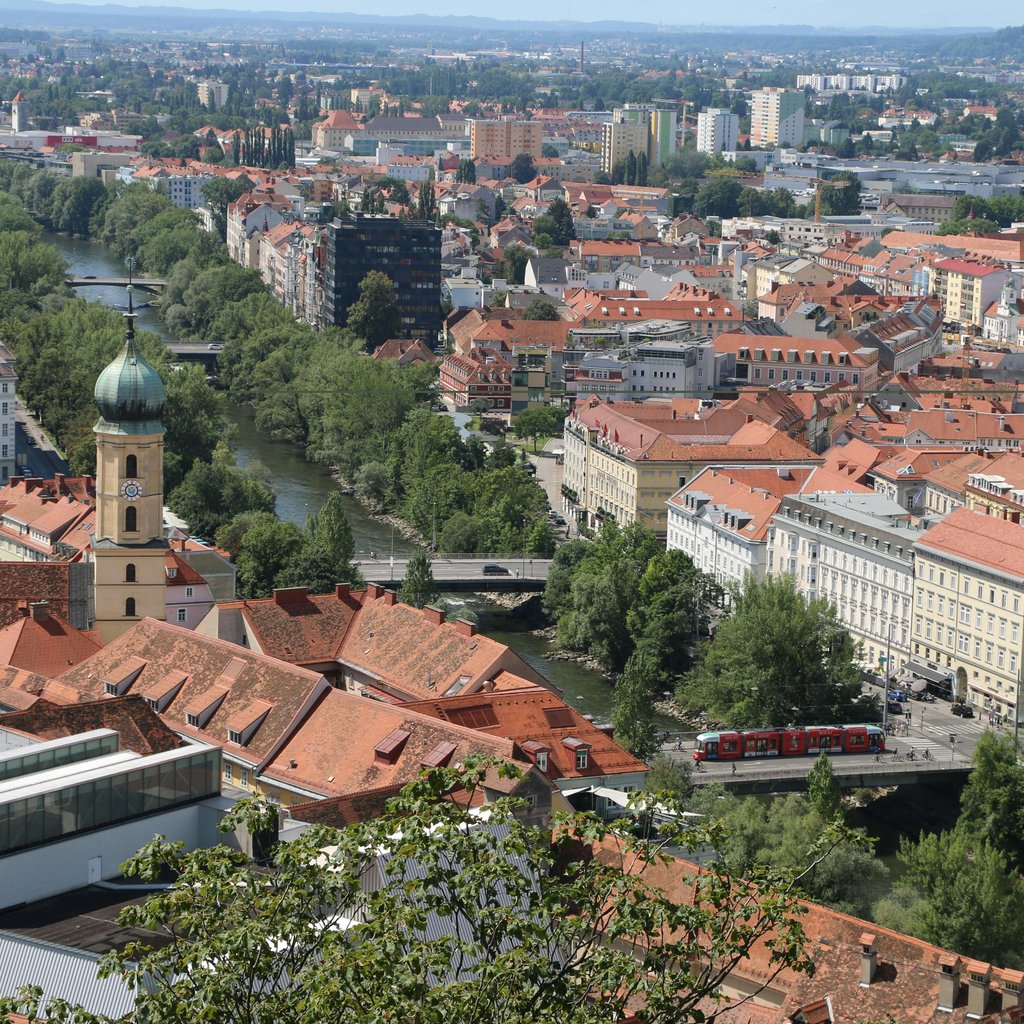 A scenic aerial view of Graz, Austria featuring red rooftops, the Mur River, and lush greenery.