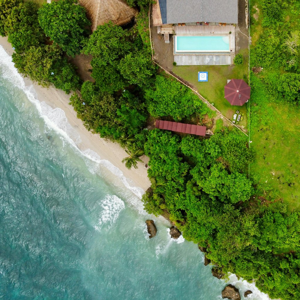 Stunning aerial shot of a tropical beachfront villa with lush greenery and a pool overlooking the ocean.