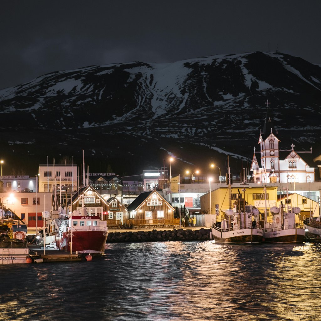 Seafront of brightly lit Husavik harbor late at night in Iceland with sailboats at quayside and dark snow covered mountain in background