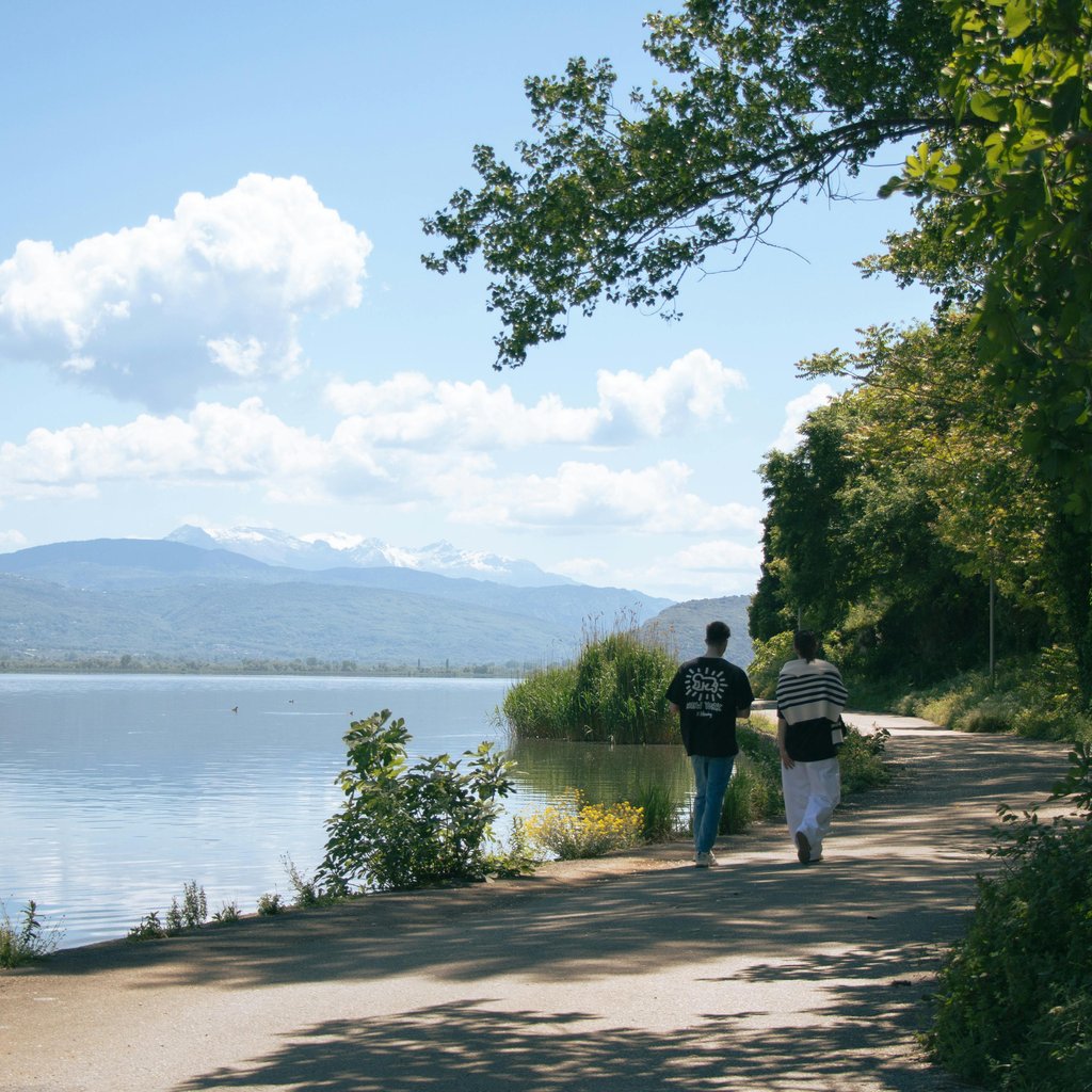 A scenic view of a couple walking along Ioannina lake in Greece under a clear blue sky.