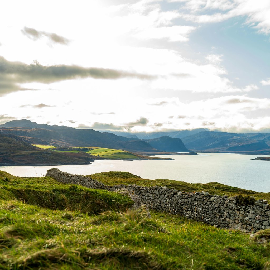 Breathtaking view of Highlands with stone wall, calm lake, and distant hills under a bright sky.
