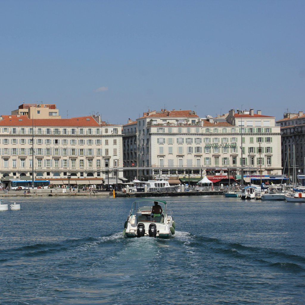 View of Marseille harbor featuring a speedboat and historic buildings on a sunny day, capturing the essence of Mediterranean coastal living.