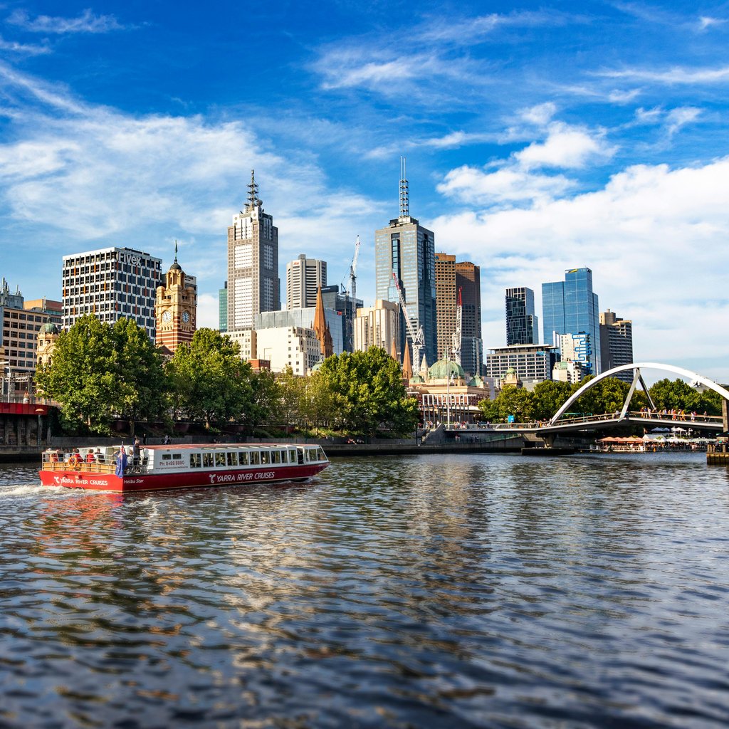 A scenic view of Melbourne's modern skyline along the Yarra River featuring a vibrant cruise boat.