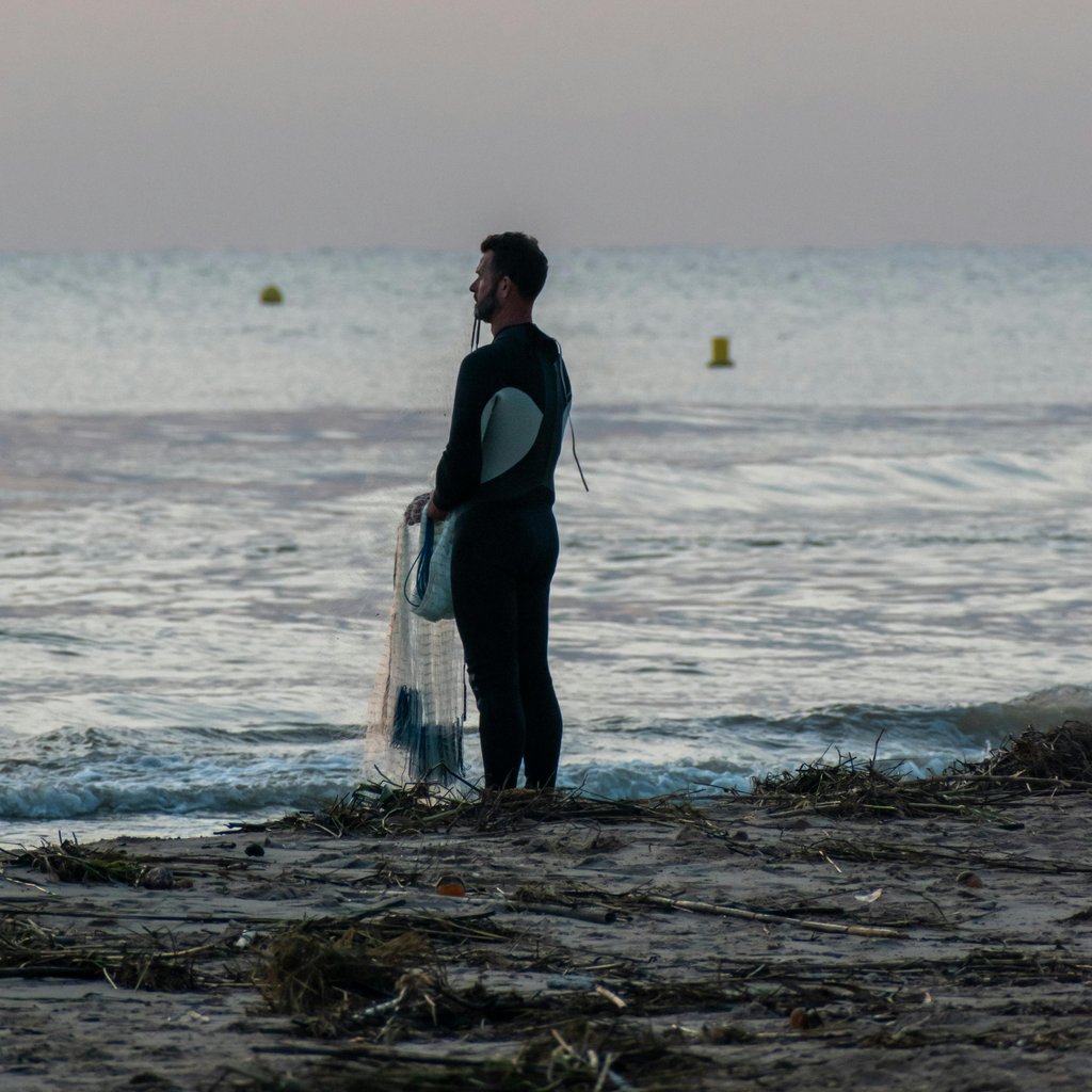 A lone surfer stands on a beach at dusk, holding a surfboard and gazing at the calm sea.