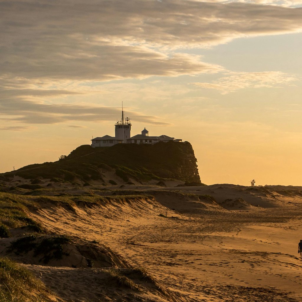 Nobby's Beach at sunset featuring a lighthouse and distant ocean vessel.