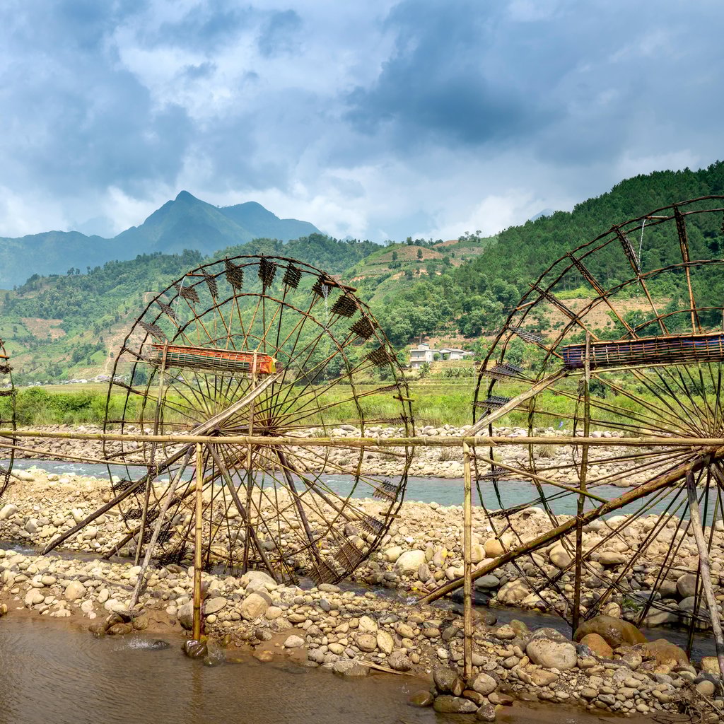Row of water wheels between bamboo fence and green mounts under cloudy sky in foggy weather