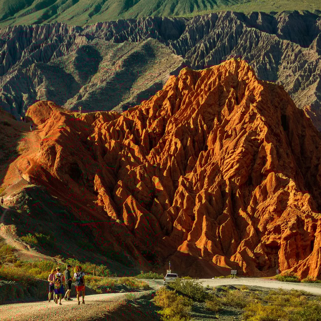Explorers trek through the vivid red hills of Purmamarca, Argentina, under a bright sky.