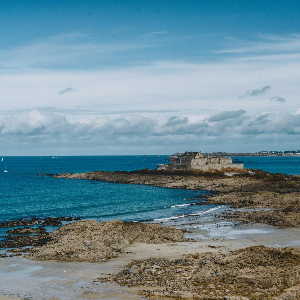 Breathtaking view of a historic fortress on the rocky coastline of Saint-Malo, France under a vibrant blue sky.
