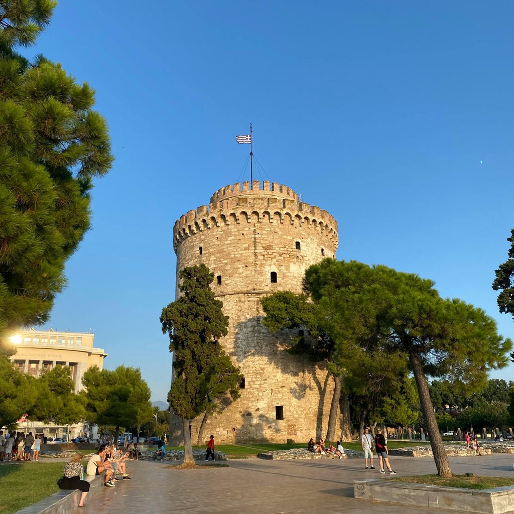 View of the iconic White Tower in Thessaloniki, Greece, under clear blue skies with surrounding trees.