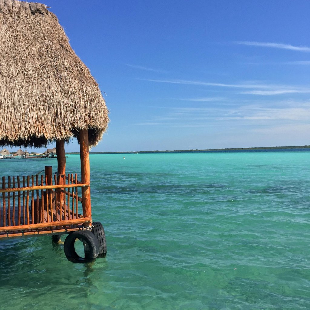 A picturesque view of Bacalar Lagoon in Mexico with a traditional thatched hut overlooking turquoise waters under a clear blue sky.