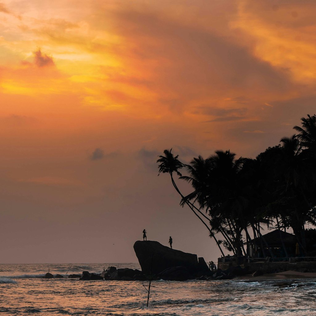 Dramatic sunset silhouette of two figures on a rocky shore at Unawatuna Beach, Sri Lanka.