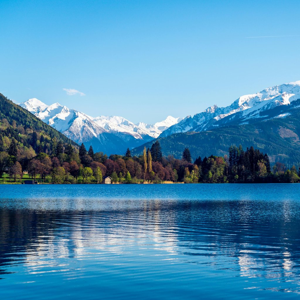 Snow-covered peaks mirrored in a tranquil lake under a clear blue sky, offering a breathtaking view of nature's beauty.